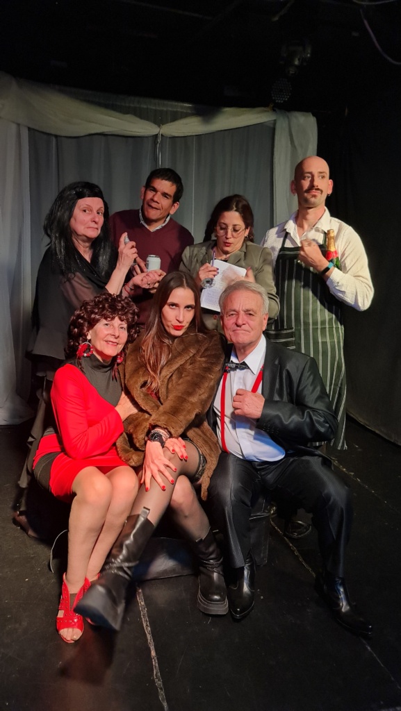 Siete personas posan en un escenario teatral con cortinas blancas de fondo. En primer plano, tres personas sentadas: una mujer mayor de rojo sonriente, una mujer joven con abrigo de piel y mirada seria, y un hombre mayor con traje y tirantes rojos. Detrás, cuatro personas de pie interpretando distintos personajes: una mujer de negro con peluca, un hombre sonriente, una mujer con gafas tomando notas, y un camarero con delantal y botella de champán.