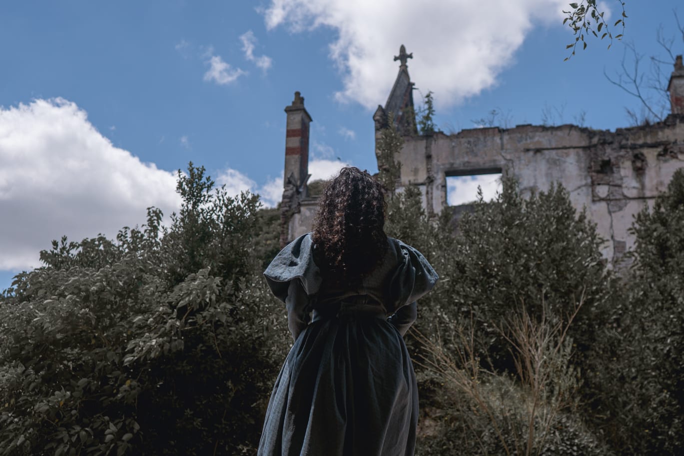 Se observa una mujer de espalda con vestido antiguo gris y delante de ella un castillo en ruinas
